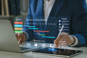 Person in blue suit in front of laptop with stylus in hand writing on tablet and project management screen superimposed