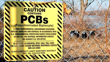 Yellow sign with "Caution Contains PCBs" on a fence with hazardous waste behind it